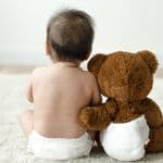 Photo from behind of a teddy bear in nappy with arm around human baby