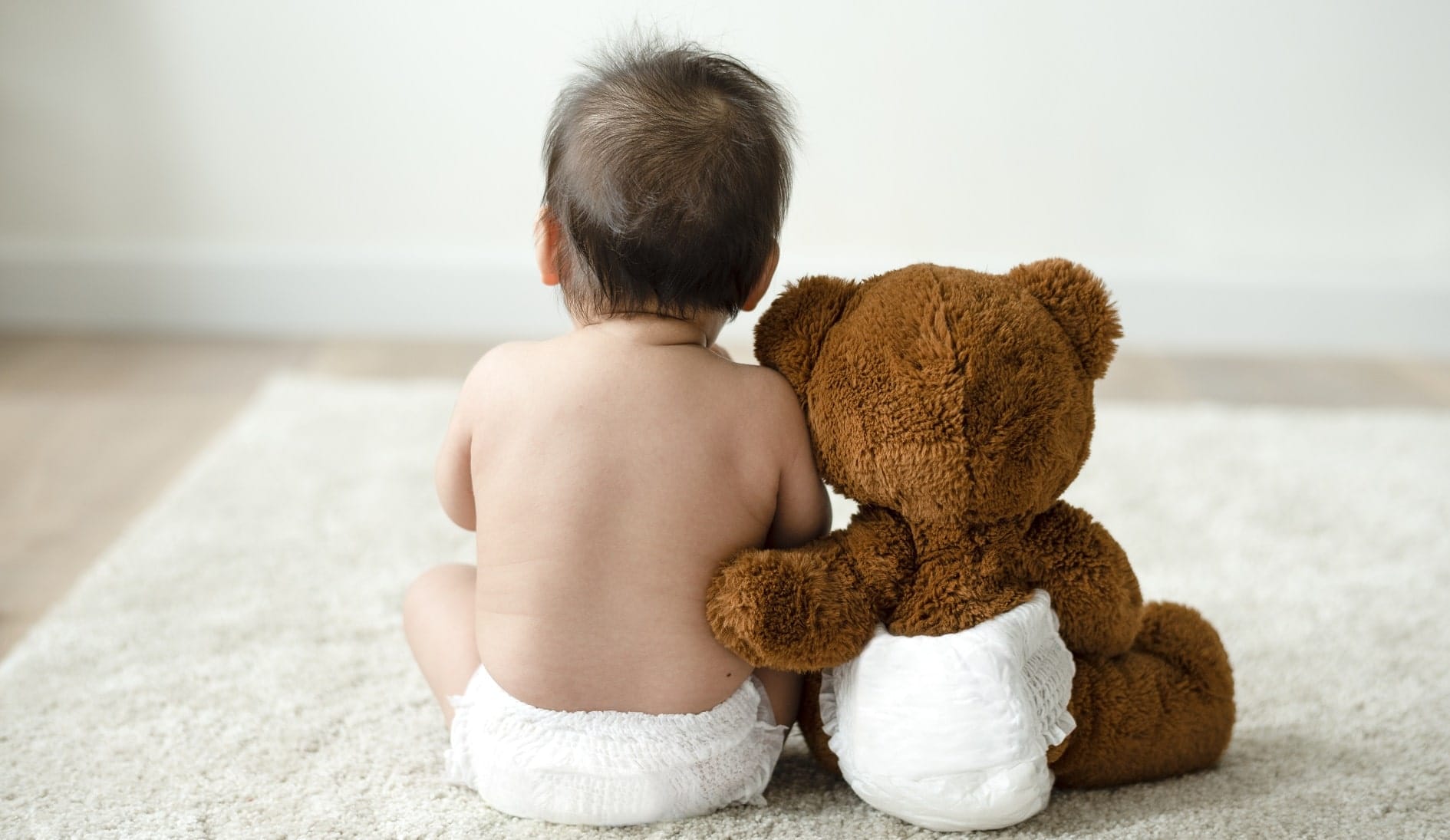 Photo from behind of a teddy bear in nappy with arm around human baby